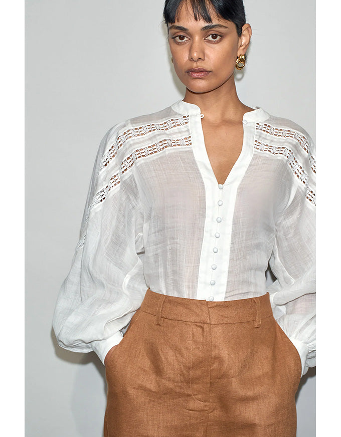 Woman wearing a white blouse with lace details and brown pants against a plain background