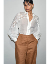 Woman wearing a white blouse with sheer sleeves and brown pants against a gray background