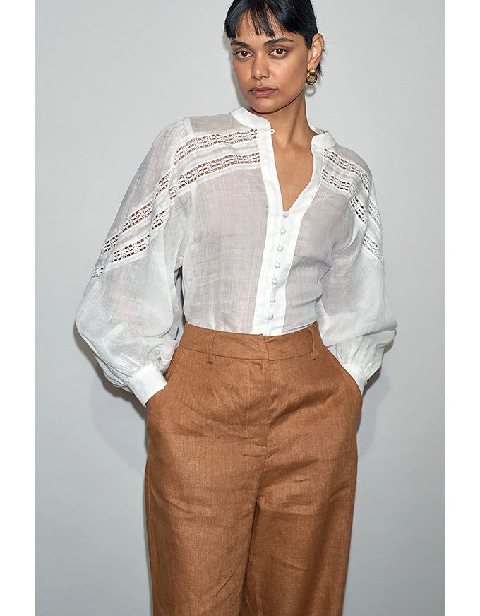 Woman wearing a white blouse with sheer sleeves and brown pants against a gray background