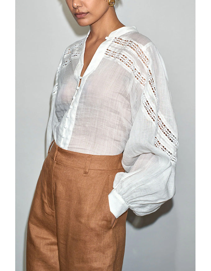 Person wearing a sheer white blouse with lace details and brown pants against a plain background