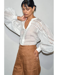 Woman wearing a white blouse with sheer sleeves and brown pants against a plain background