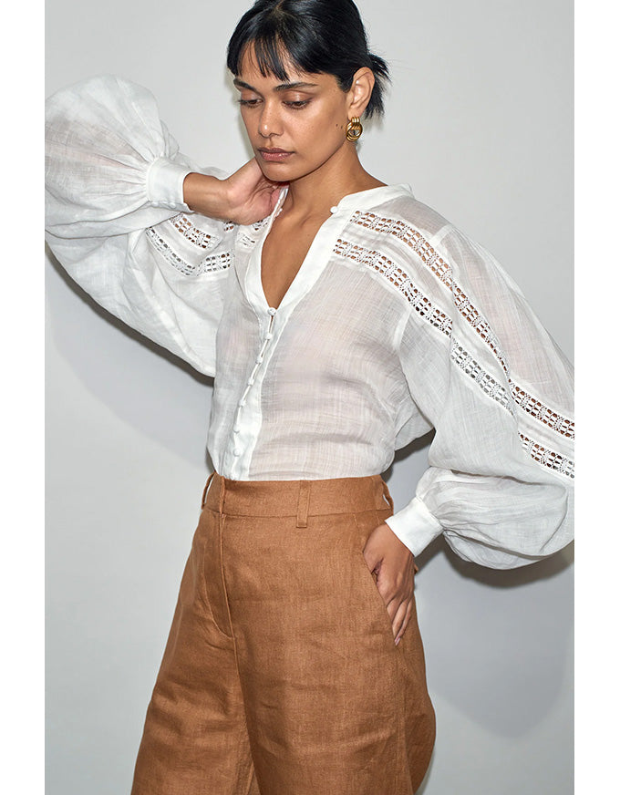 Woman wearing a white blouse with sheer sleeves and brown pants against a plain background