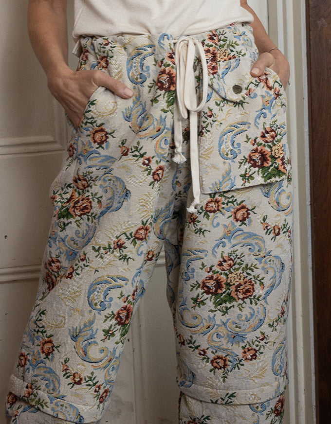 A person wearing wide-leg cropped pants with a cream base and an ornate blue, pink, and green floral scroll pattern, paired with a white drawstring waist and cargo-style pockets.