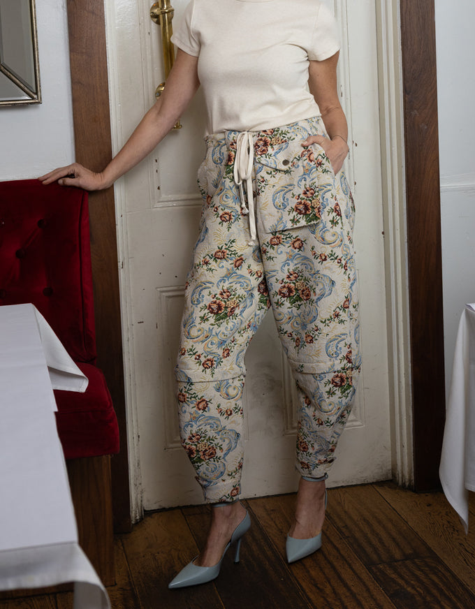 A person wearing patterned drawstring pants with a cream top and light blue heels, standing indoors near a white door and wooden floor.