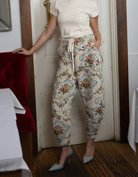 A person wearing patterned drawstring pants with a cream top and light blue heels, standing indoors near a white door and wooden floor.