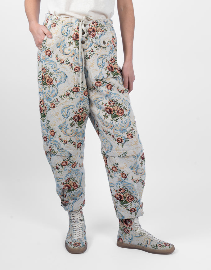 A person wearing loose, tapered pants with an allover floral and scroll pattern in muted blue, red, green, and beige tones, paired with matching lace-up boots against a plain light gray studio background.