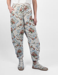 A person wearing loose, tapered pants with an allover floral and scroll pattern in muted blue, red, green, and beige tones, paired with matching lace-up boots against a plain light gray studio background.