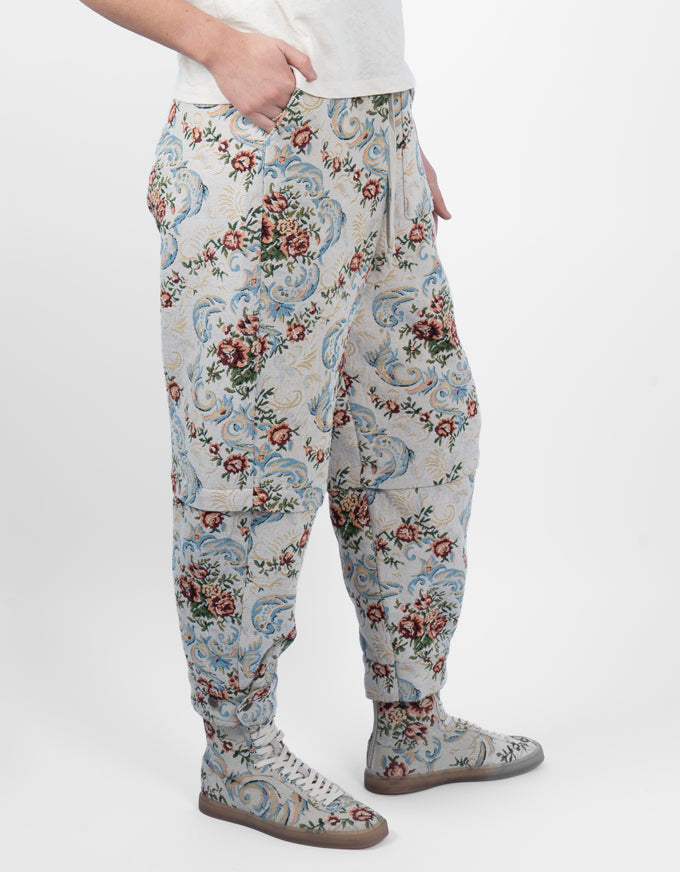 A person wearing loose-fit patterned pants with a cream base and blue, red, and green floral and scroll motifs, paired with matching lace-up boots against a light gray studio background.