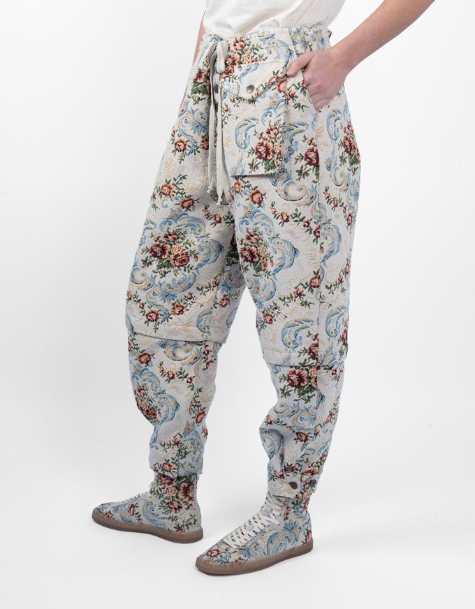A person wearing loose, high-waisted jogger-style pants with an allover floral and paisley print in muted blue, red, green, and beige tones, paired with matching sneakers, against a clean white studio background.