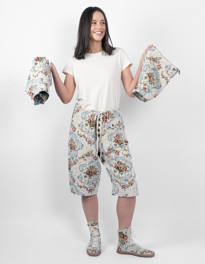 A woman stands against a light gray studio background wearing a white t-shirt, cream cropped pants with a blue and red floral print, and matching floral socks. She holds up matching floral pouches or small fabric items in both hands.
