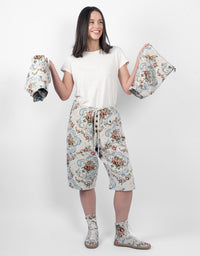A woman stands against a light gray studio background wearing a white t-shirt, cream cropped pants with a blue and red floral print, and matching floral socks. She holds up matching floral pouches or small fabric items in both hands.