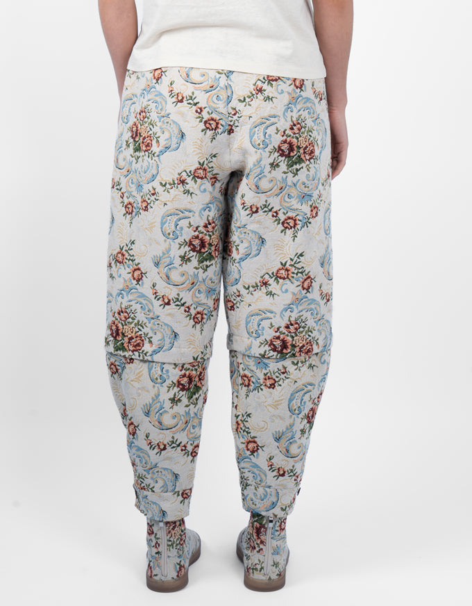A person shown from the back wearing loose, tapered pants with a dense floral and scroll pattern in blue, red, and green on a light beige base, paired with a white top and gray boots against a plain white studio background.
