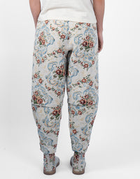 A person shown from the back wearing loose, tapered pants with a dense floral and scroll pattern in blue, red, and green on a light beige base, paired with a white top and gray boots against a plain white studio background.