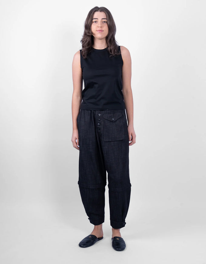 A woman stands against a light gray studio background wearing a black sleeveless top and dark wide-leg pants with a button front and large front pockets, along with dark slip-on shoes.