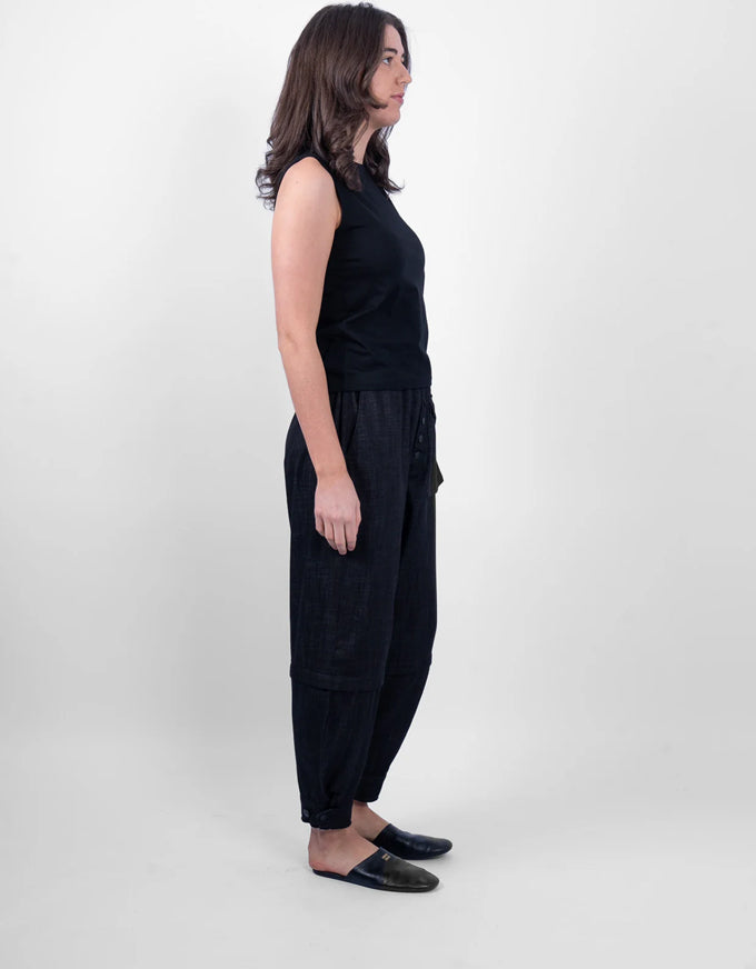 A woman stands in profile wearing a black sleeveless top and loose black pants with dark slip-on shoes, photographed against a plain light gray studio background.