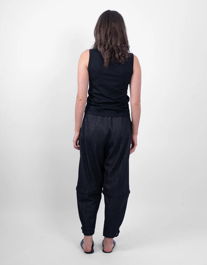 A woman viewed from behind wearing a black sleeveless top and dark charcoal gray relaxed-fit pants, standing against a plain light gray studio background.