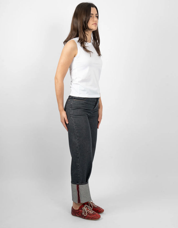 A woman stands in profile wearing a white sleeveless top, dark gray slim-fit jeans with rolled cuffs showing a gray lining, and red lace-up shoes, against a light gray studio background.