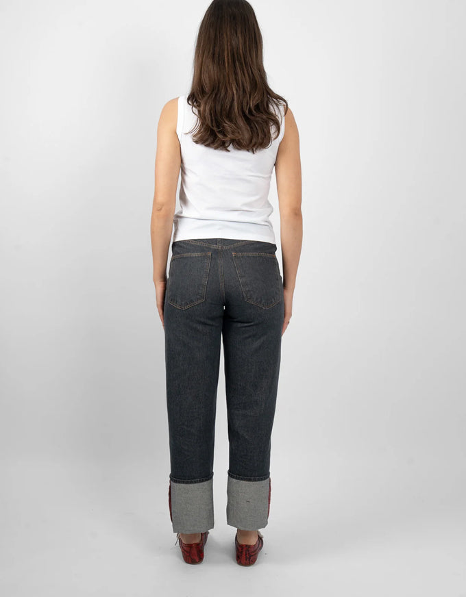A woman viewed from behind wearing dark gray cuffed jeans, a white sleeveless top, and brown shoes against a light gray studio background.