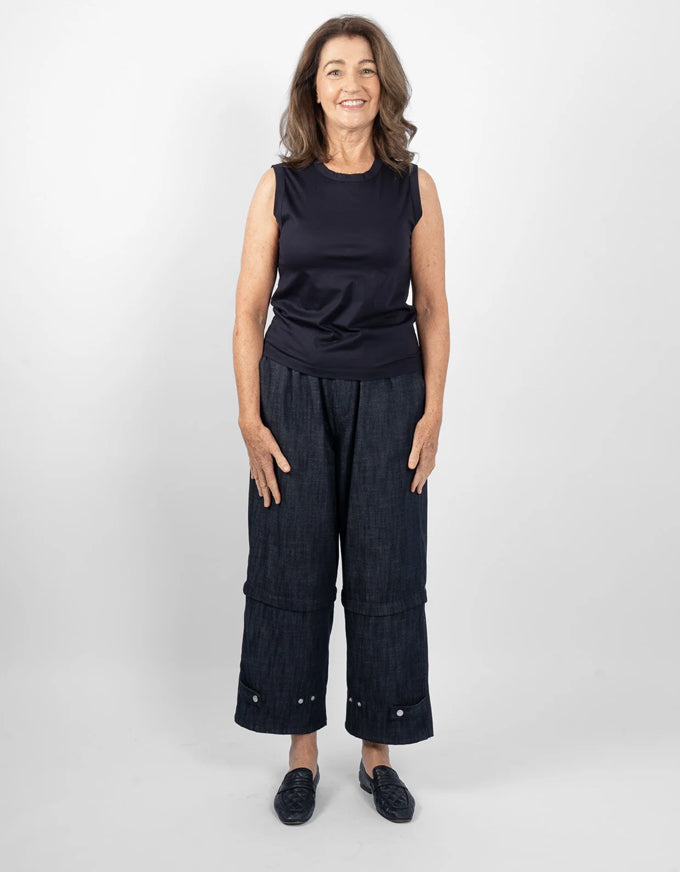 A woman modeling a sleeveless dark navy top and dark wide-leg denim pants with rolled cuffs and metal rivet details, standing against a light gray studio background.