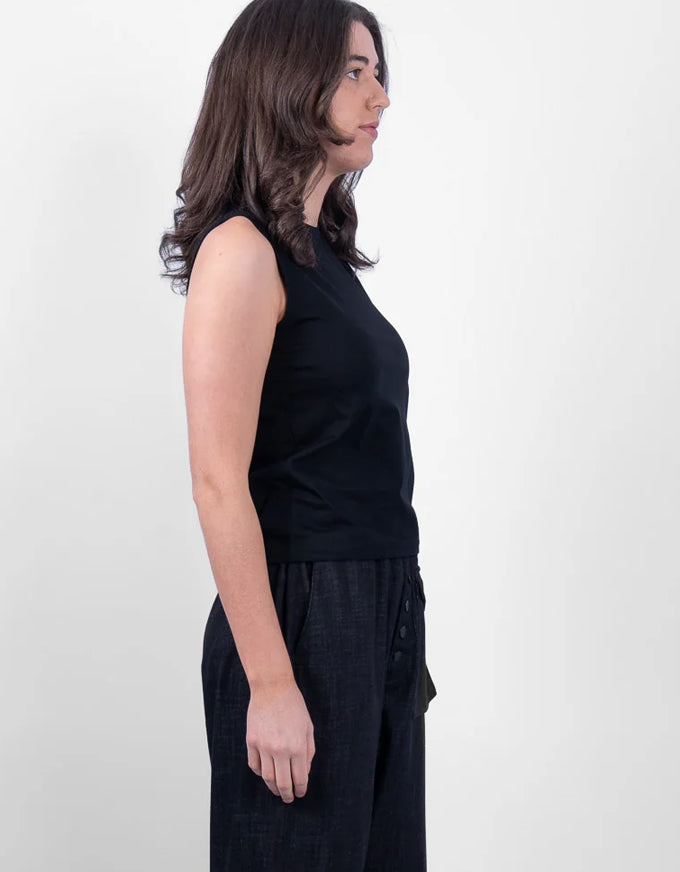 A woman shown in profile wearing a sleeveless black top and dark wide-leg pants against a plain light gray studio background.