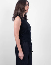 A woman shown in profile wearing a sleeveless black top and dark wide-leg pants against a plain light gray studio background.