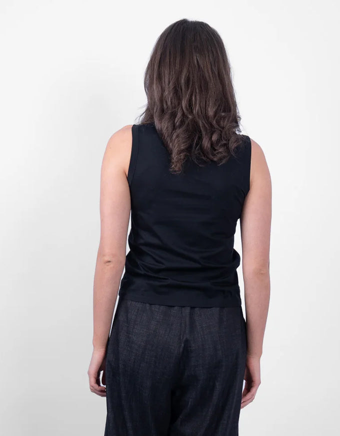 A woman viewed from behind wearing a black sleeveless top and dark textured pants against a plain light background.