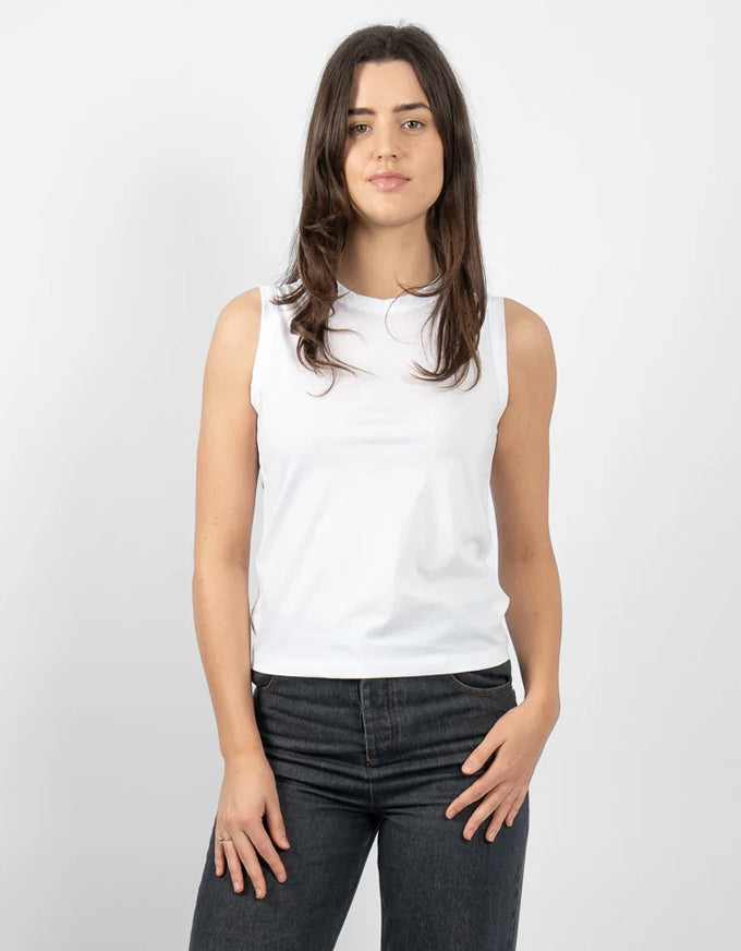 A woman modeling a white sleeveless crewneck tank top paired with dark gray jeans, standing against a light gray studio background.