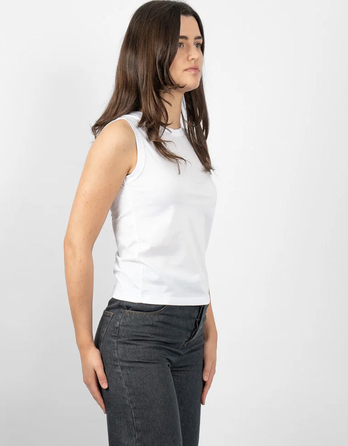 A woman wearing a plain white sleeveless crop top and dark gray jeans, shown from a side profile against a light gray studio background.