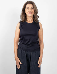 A smiling woman with shoulder-length brown hair stands against a light gray studio background, wearing a sleeveless dark navy top and dark wide-leg pants.