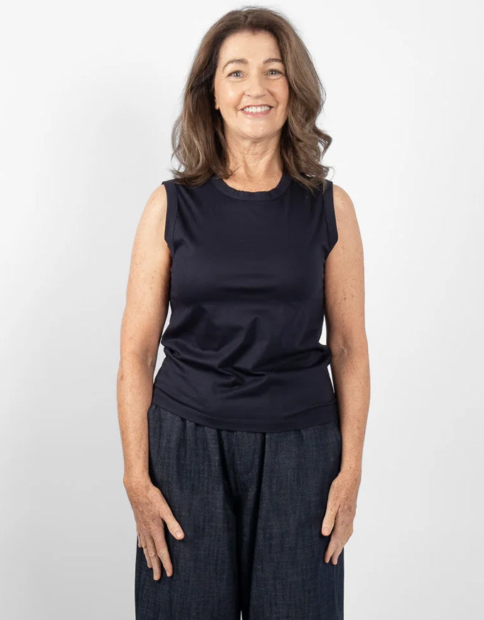 A smiling woman with shoulder-length brown hair stands against a light gray studio background, wearing a sleeveless dark navy top and dark wide-leg pants.
