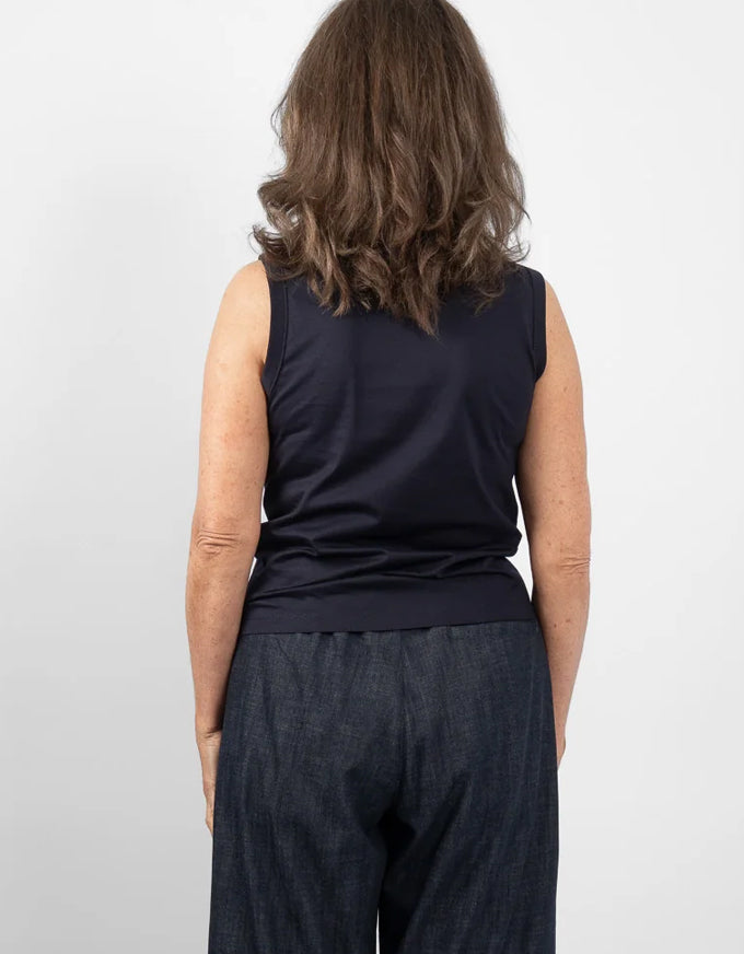 A woman viewed from behind wearing a dark navy sleeveless top and dark textured pants against a light gray studio background.